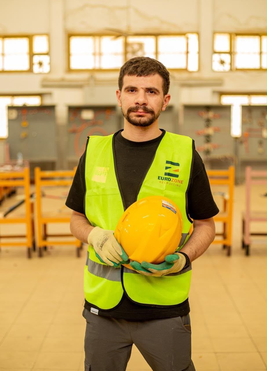A young man in a Eurozone high-visibility vest and work gloves holding a yellow hard hat in front of him, in a training hall lined with wheeled workbenches