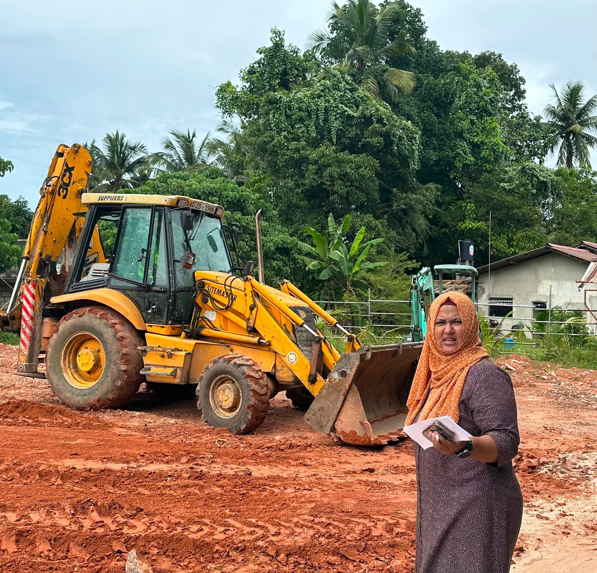 A woman holding papers on a cleared red-earth site, a yellow backhoe loader behind her and palm trees beyond