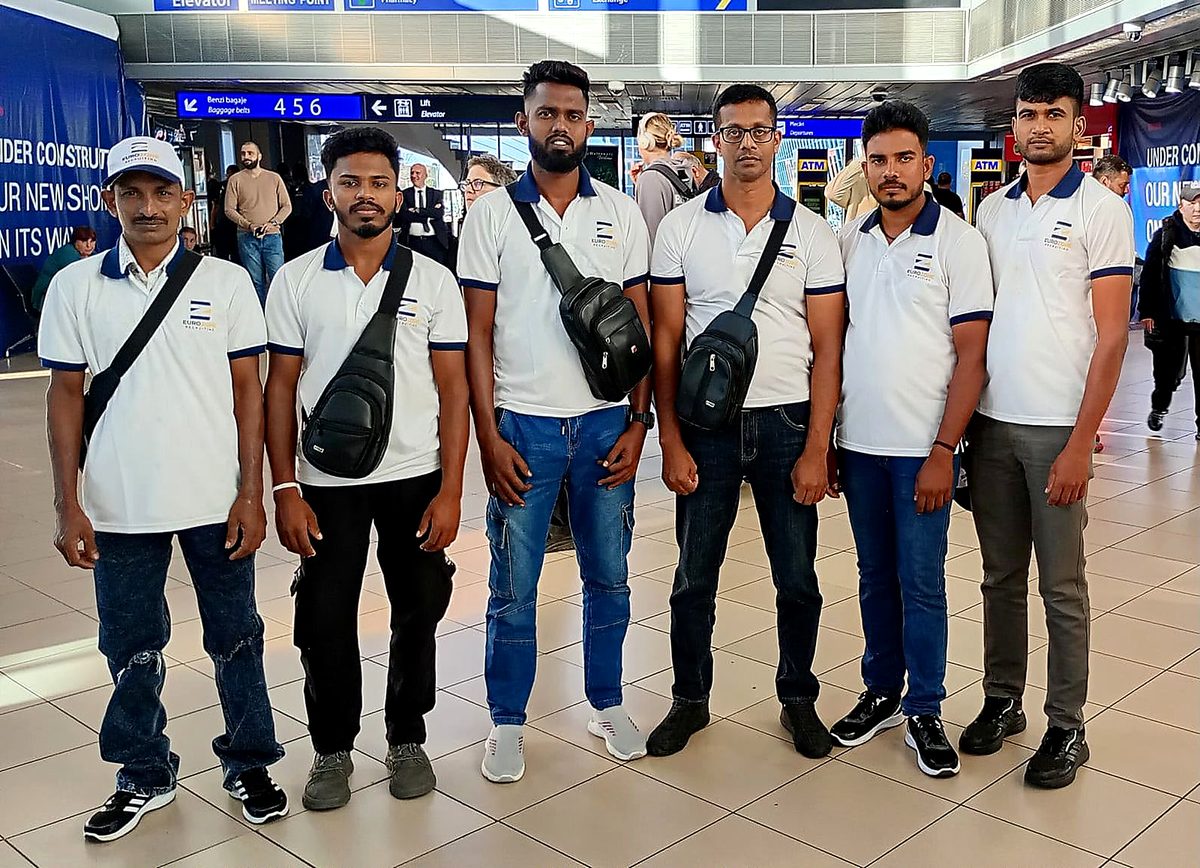 Six workers in matching Eurozone polo shirts standing in a row in an airport arrivals hall, under the baggage-belt signs