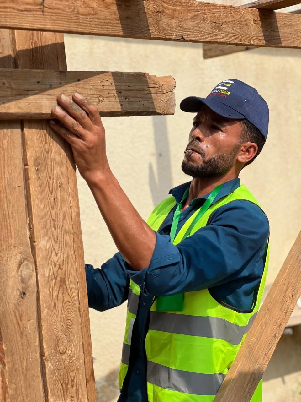 A carpenter in a navy Eurozone cap and a high-visibility vest fixing a timber beam into a wooden frame with a hammer