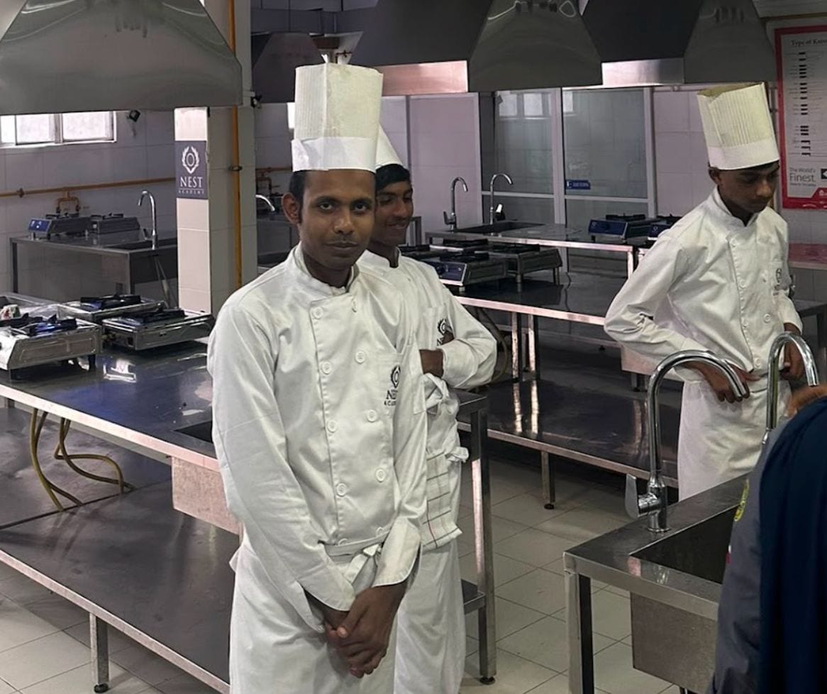 Three trainee chefs in white jackets and tall toques standing in a steel-benched training kitchen