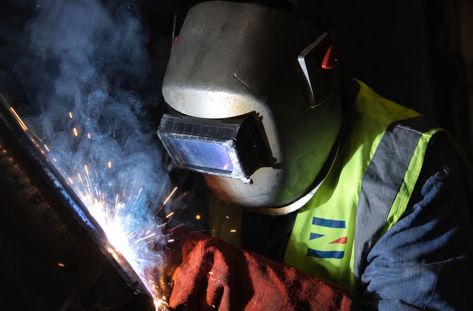 A welder in a mask and a Eurozone high-visibility vest welding a steel seam, sparks and smoke in the dark