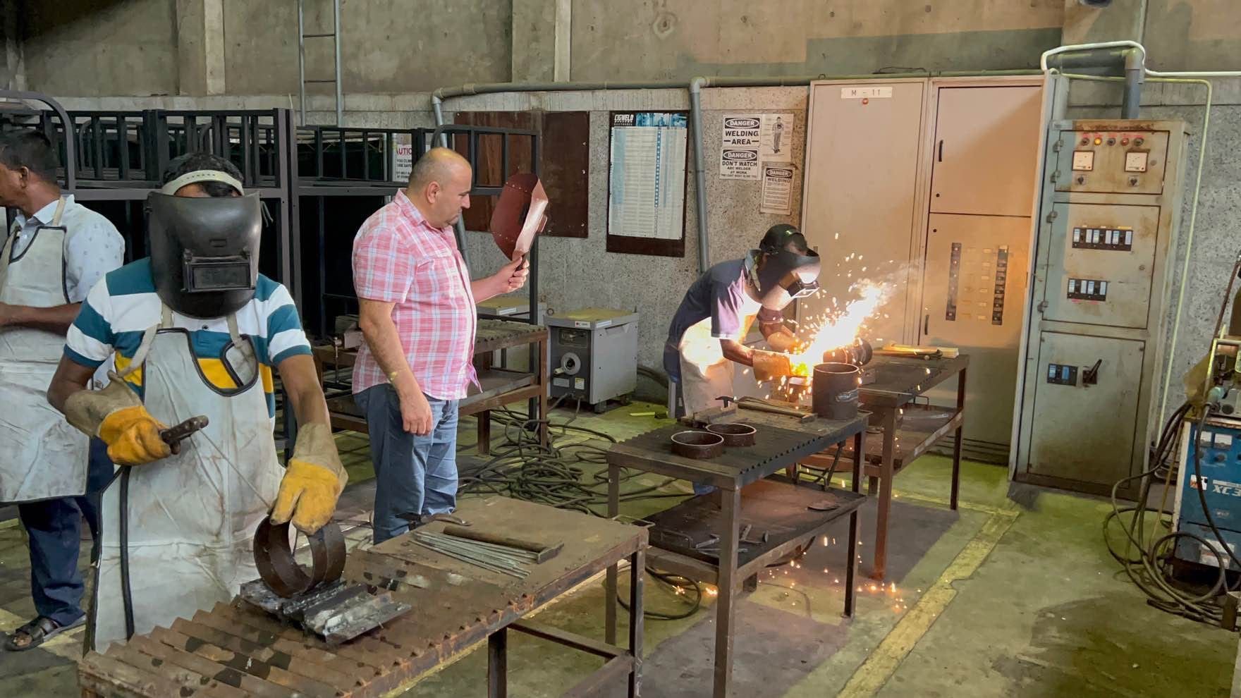 Trainees welding in a workshop, sparks flying at a bench while an instructor in a pink shirt looks on