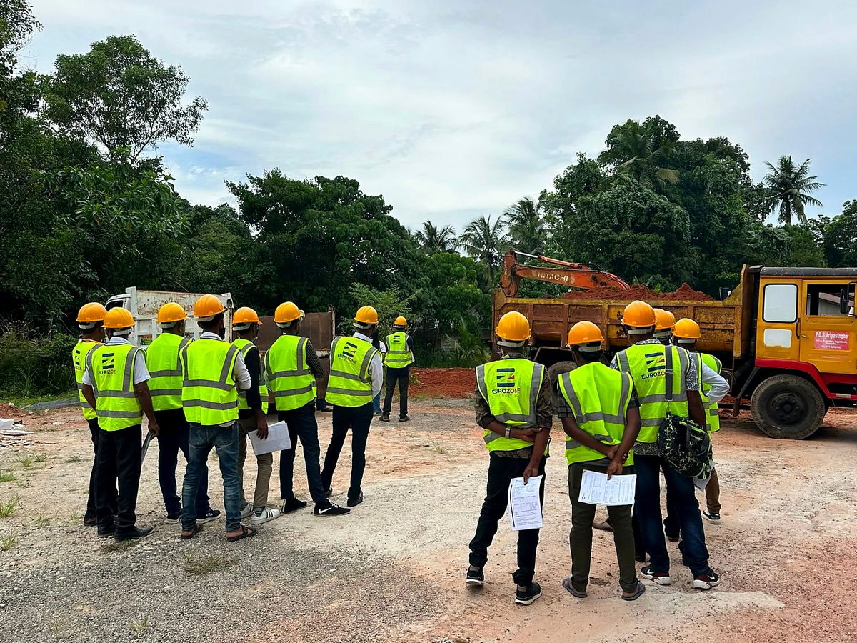 Candidates in Eurozone high-visibility vests and yellow hard hats on a site visit, papers in hand, watching an excavator load a tipper truck