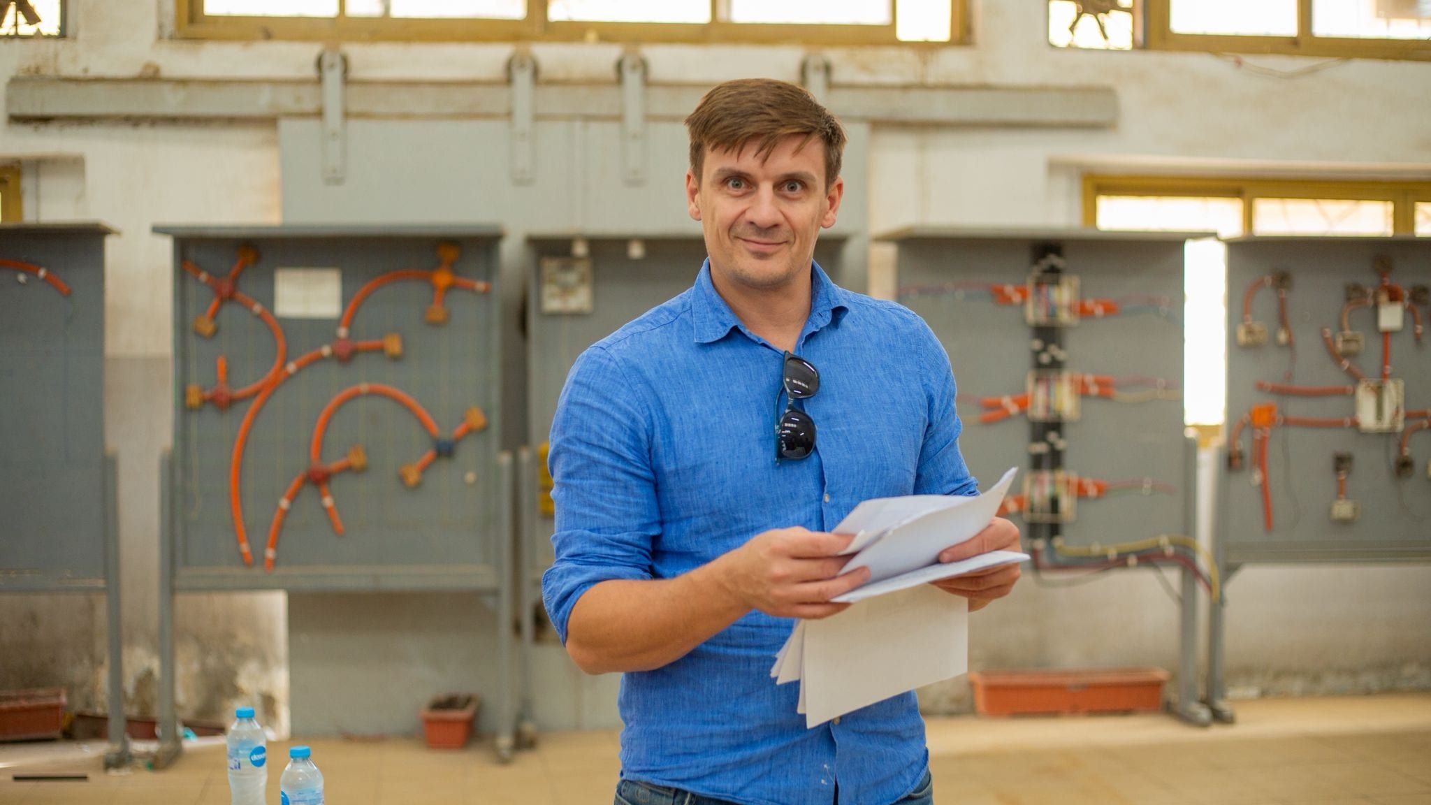 A man in a blue shirt holding a sheaf of papers, smiling at the camera in a training hall lined with electrical wiring boards.
