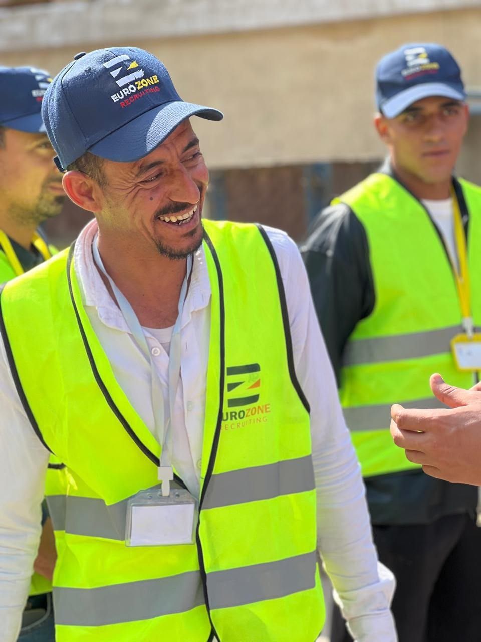 A smiling candidate in a navy Eurozone Recruiting cap and a Eurozone high-visibility vest with a name badge, talking with others in matching caps and vests