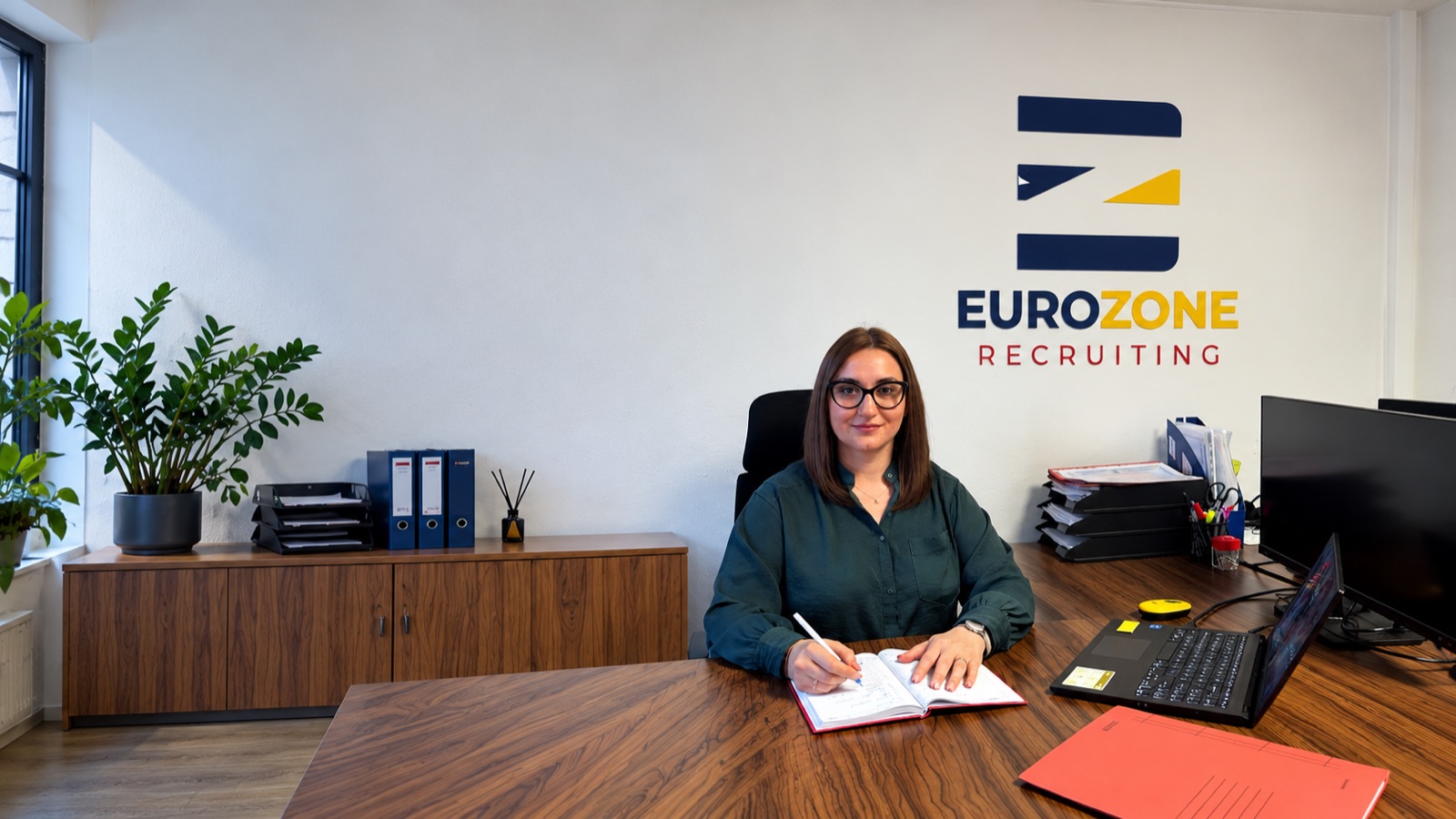 A consultant at her desk with a notebook and laptop, the Eurozone Recruiting logo on the wall behind her
