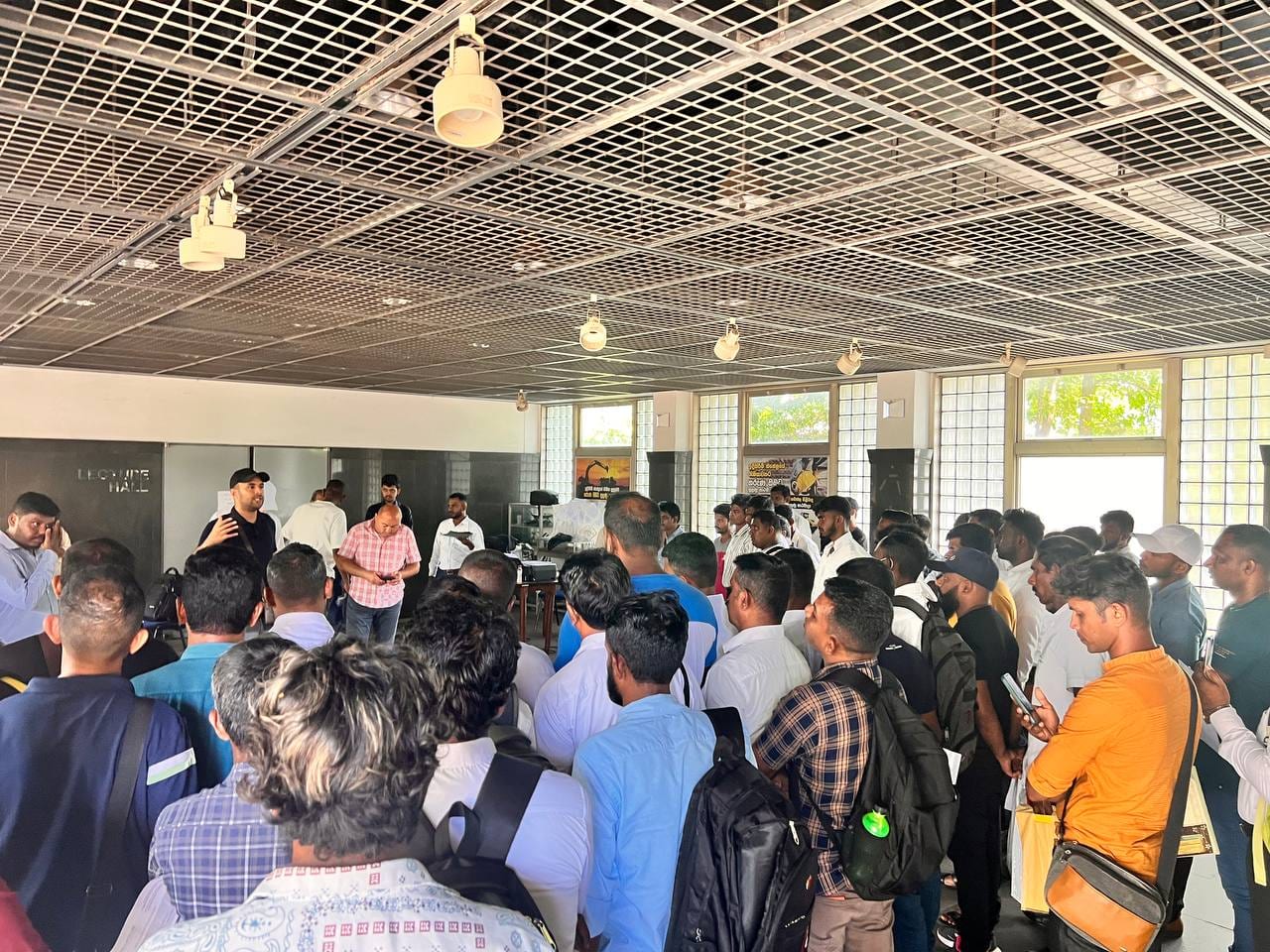 A lecture hall packed with dozens of men holding folders and backpacks, listening to a man addressing them from the front