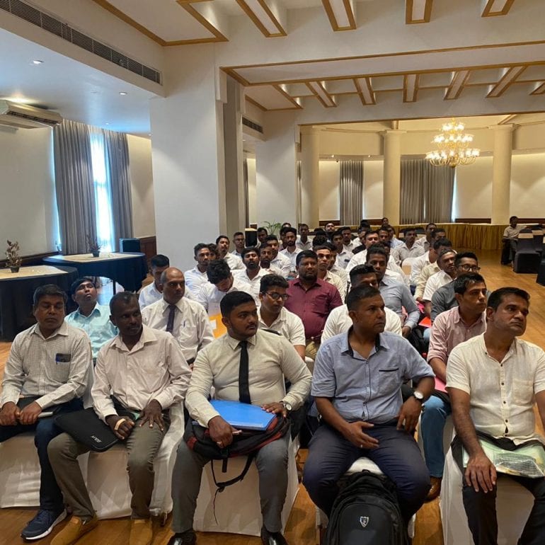 Rows of men in light shirts seated on banquet chairs in a chandeliered hotel function room, several holding blue document folders, listening to a briefing