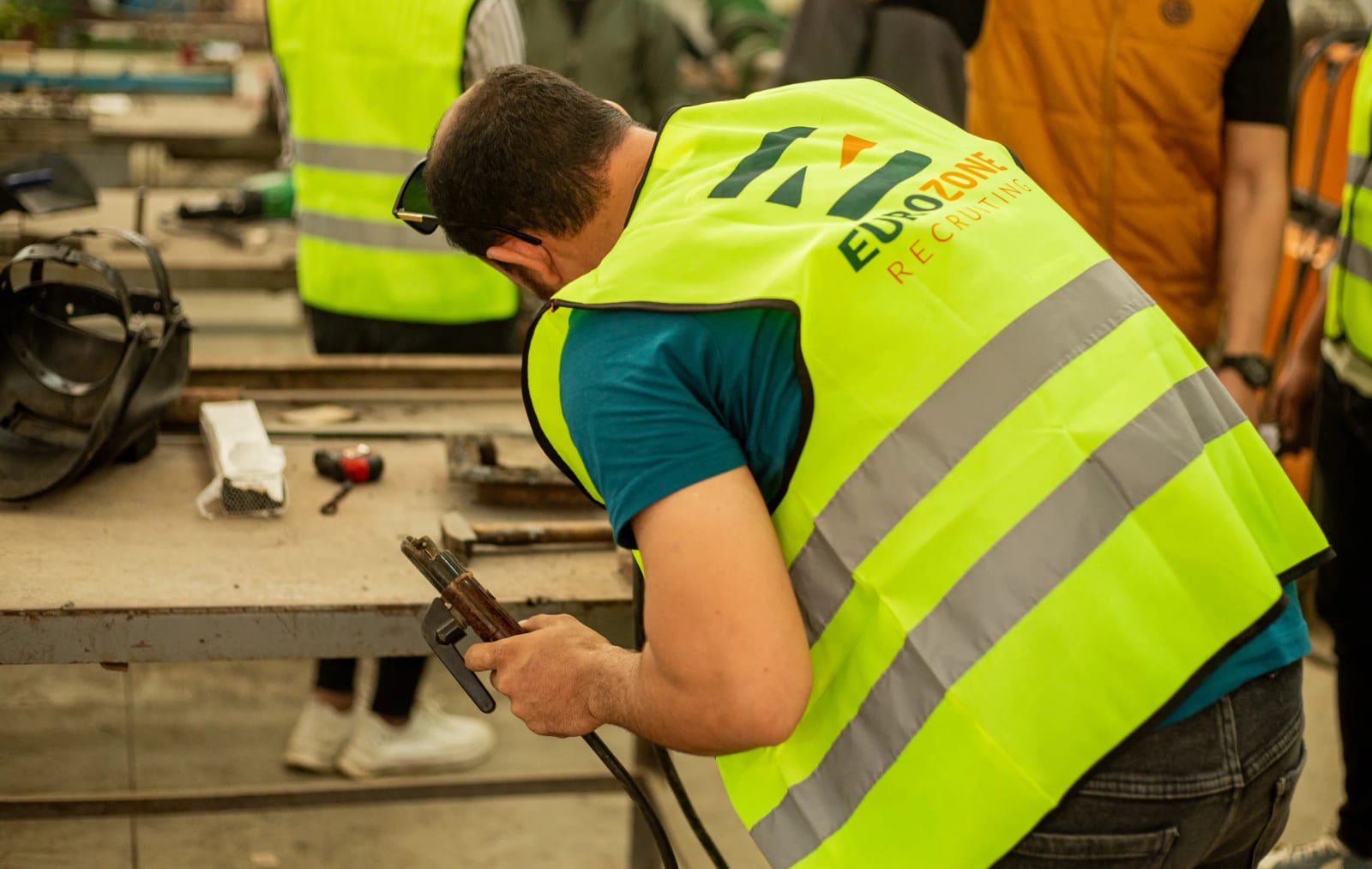 A worker in a Eurozone Recruiting hi-vis vest, seen from behind, holding a welding electrode holder at a workshop bench.