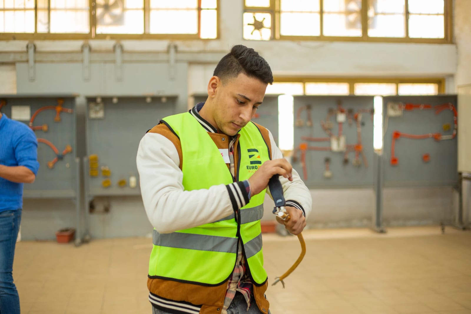 A young man in a Eurozone high-visibility vest holding a welding electrode holder on its yellow lead, in a training hall lined with wall-mounted panels
