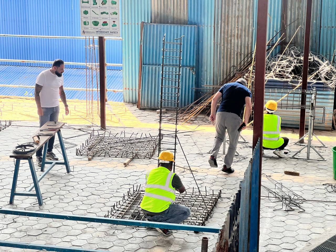 Two workers in hard hats crouched over rebar mats on a paved yard, tying the bars together