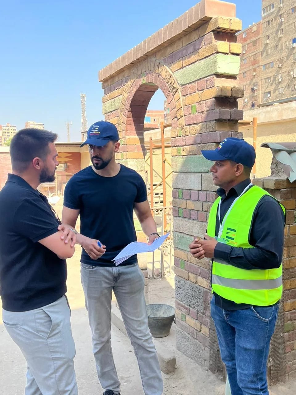 ثلاثة رجال يتحدثون بجوار قوس من الطوب قيد البناء في موقع إنشاءات، أحدهم يحمل حزمة أوراق وآخر يرتدي سترة يوروزون العاكسة